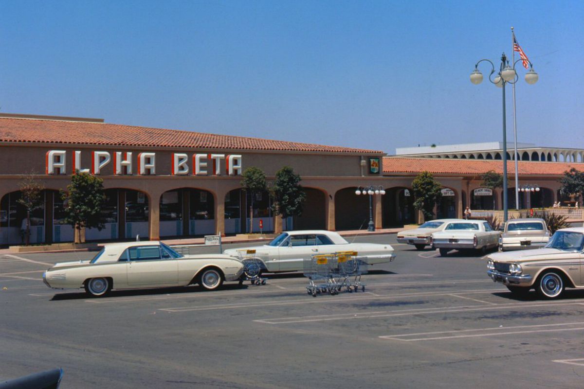 Front exterior of Alpha Beta Grocery Store, Laguna Hills, California, 1966, a few beige vintage cars are parked in the parking lot in the foreground, a blue sky in the background