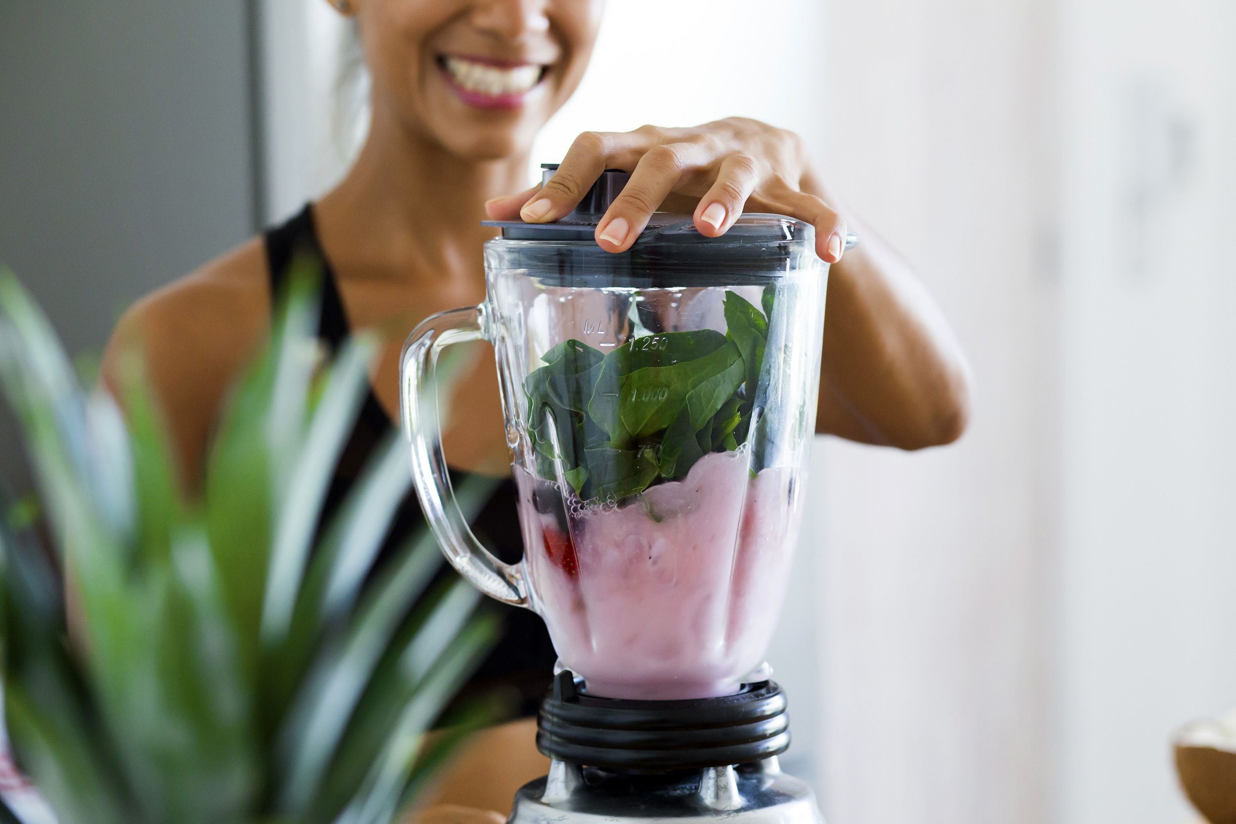 Blender filled with a light purple liquid, berries, and spinach, being blended, selective focus, by a smiling woman, blurred plant in the left foreground, dark grey and white wall blurred in the background