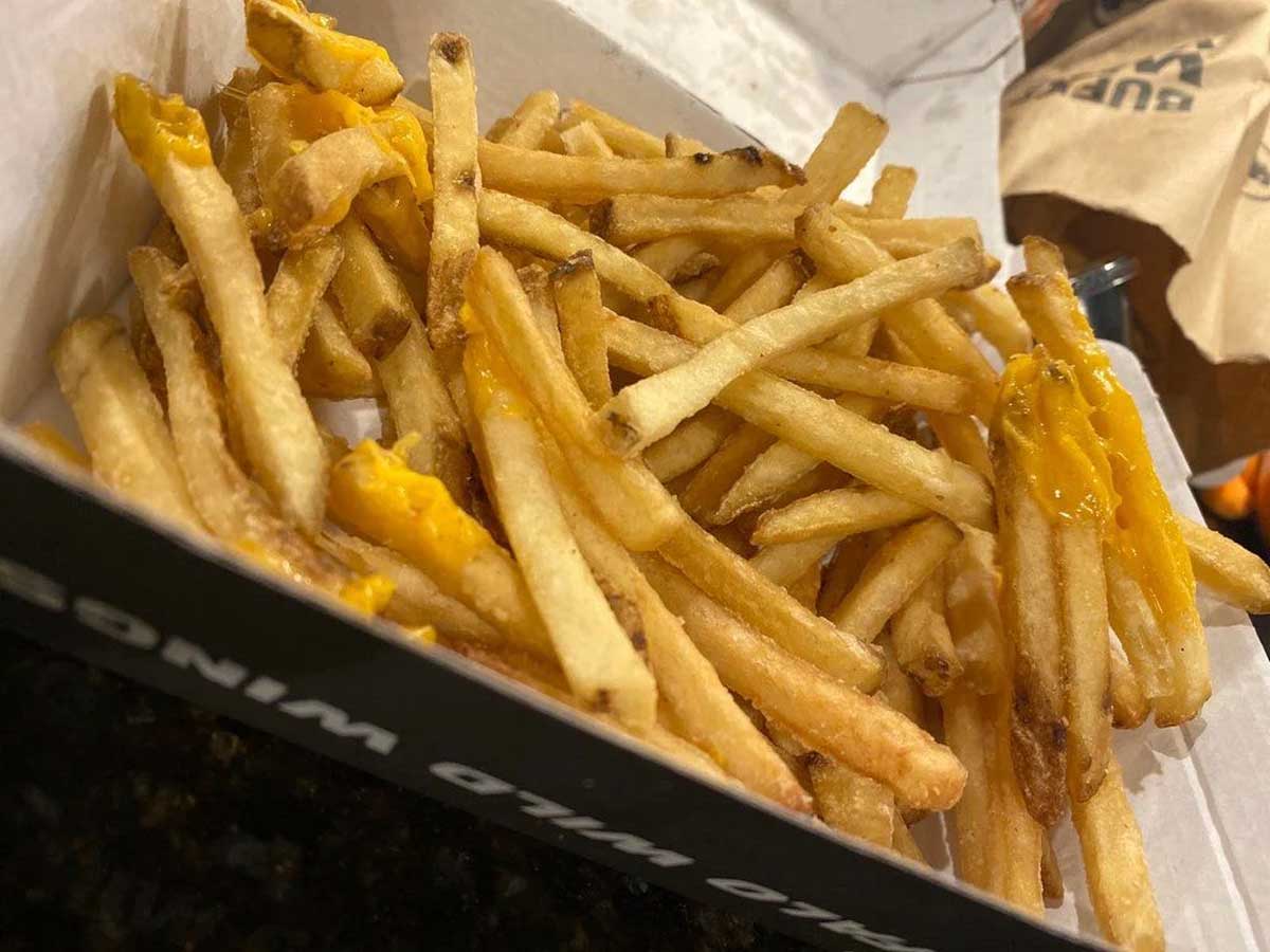 A tray of crispy beef tallow fries, with a few topped with melted cheddar cheese, served in a rectangular paper container.