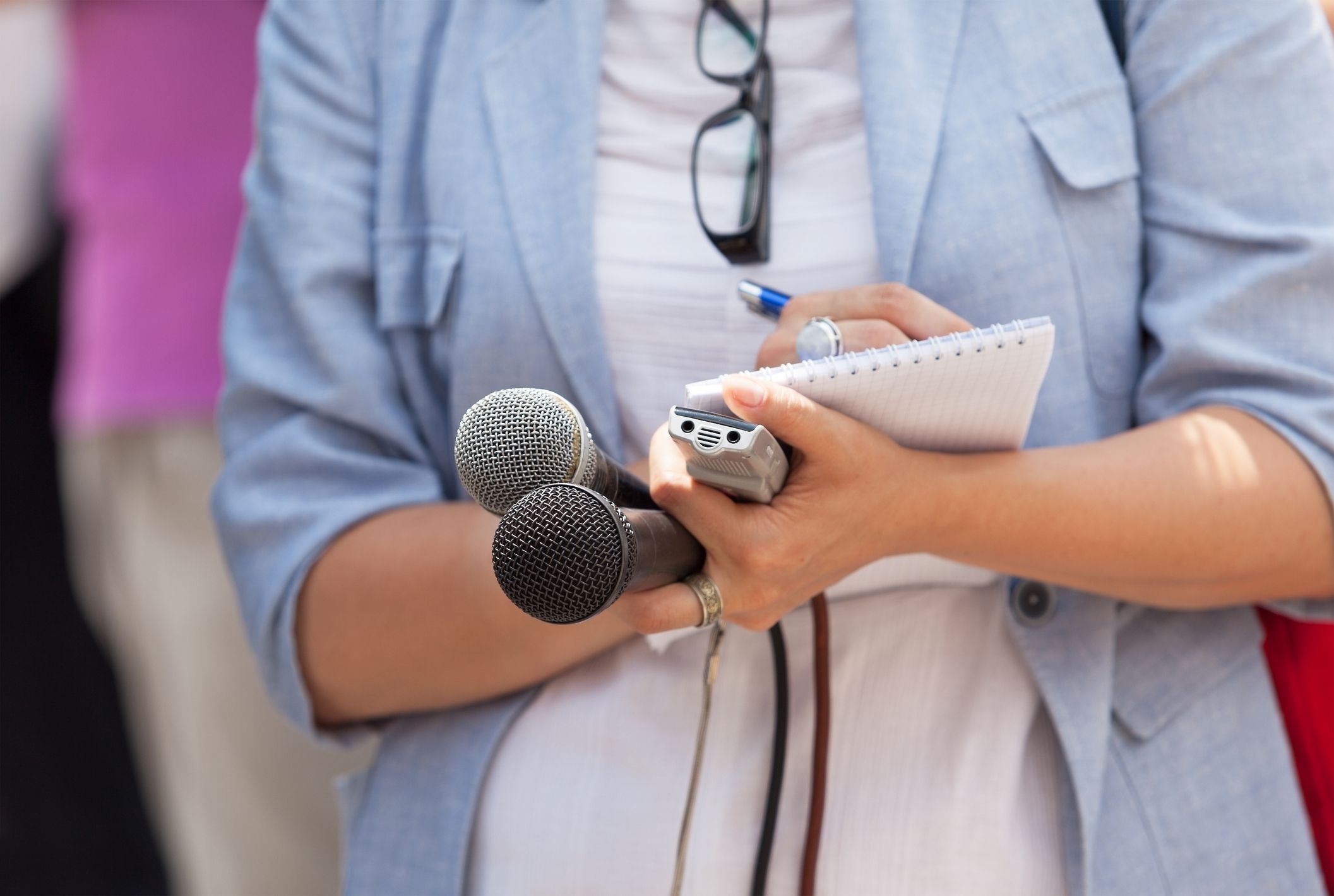 Female media reporter taking notes while on site during interview, her hands holding notebook and two microphones and a recorder, she is wearing a blue blazer and white shirt