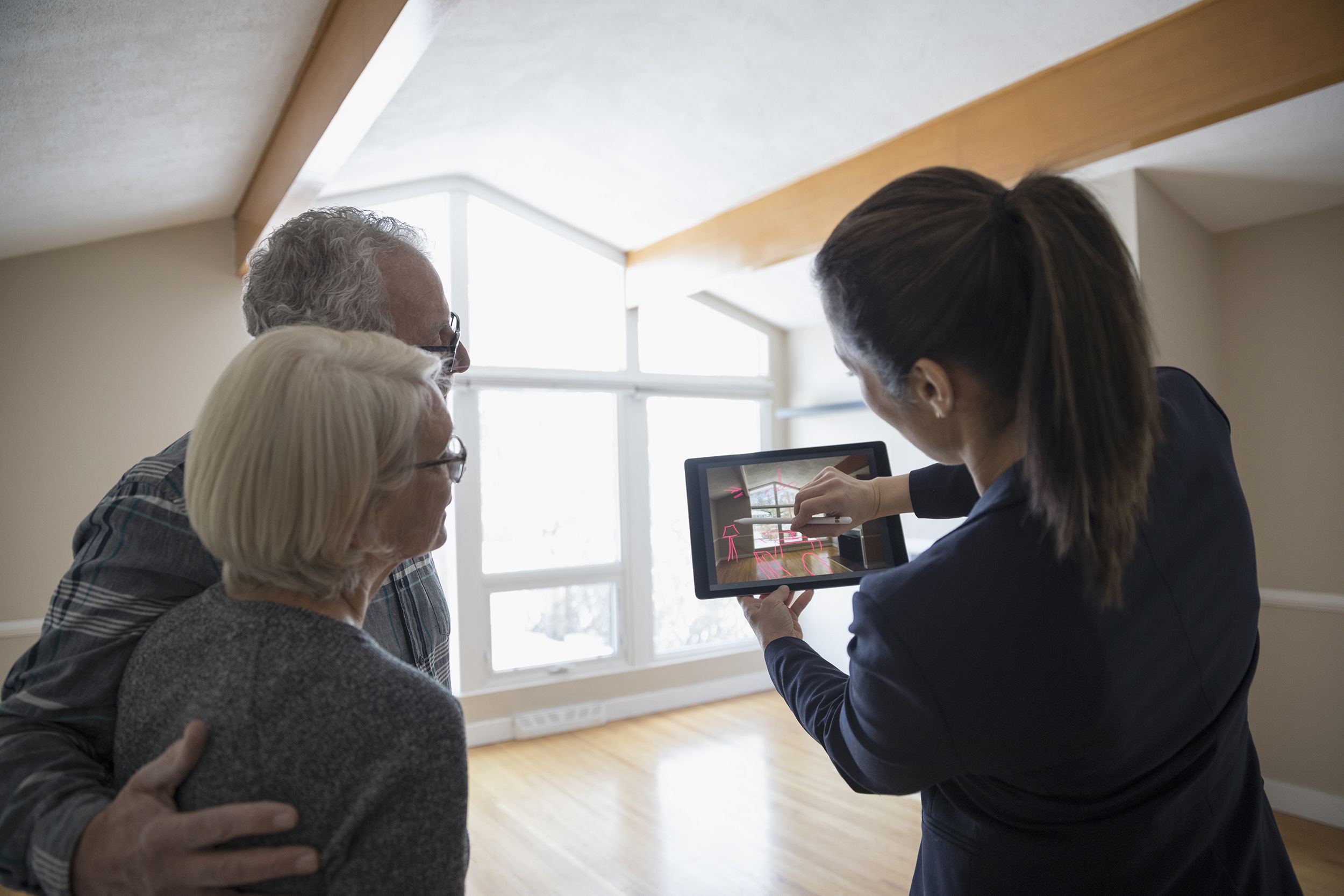 Female realtor showing senior couple living room of home for sale while using a tablet for ideas, light shining through windows of living room in the background