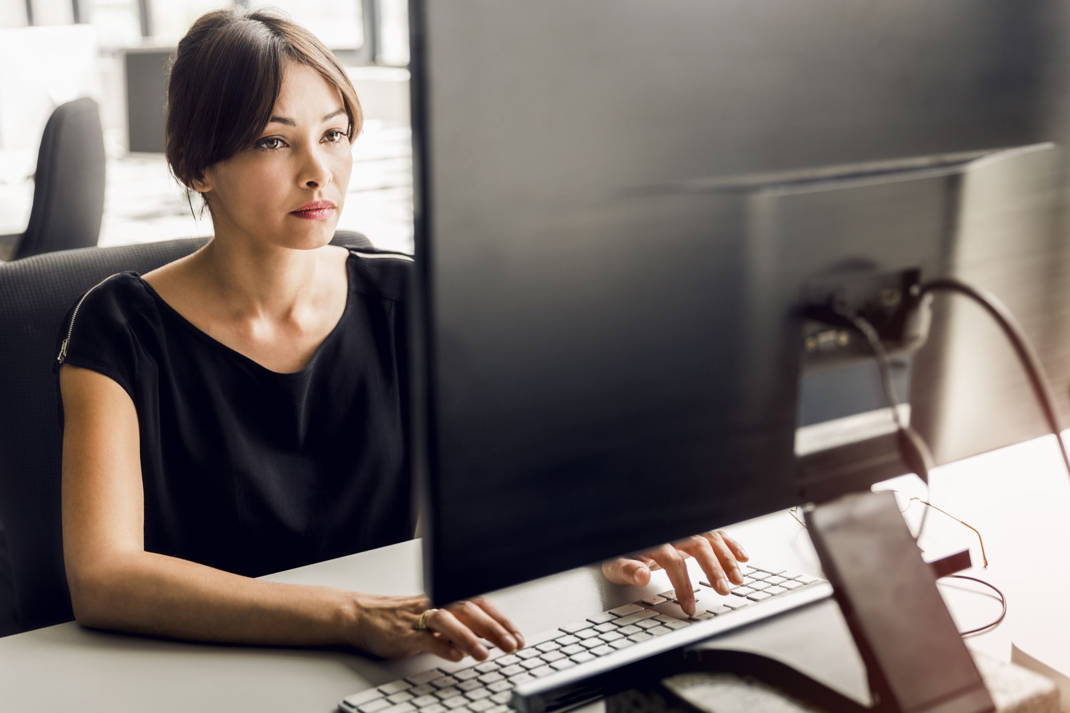 Female transcriptionist using desktop computer in office, looking at screen while typing, facing screen back of computer screen is shown blurred background of empty office