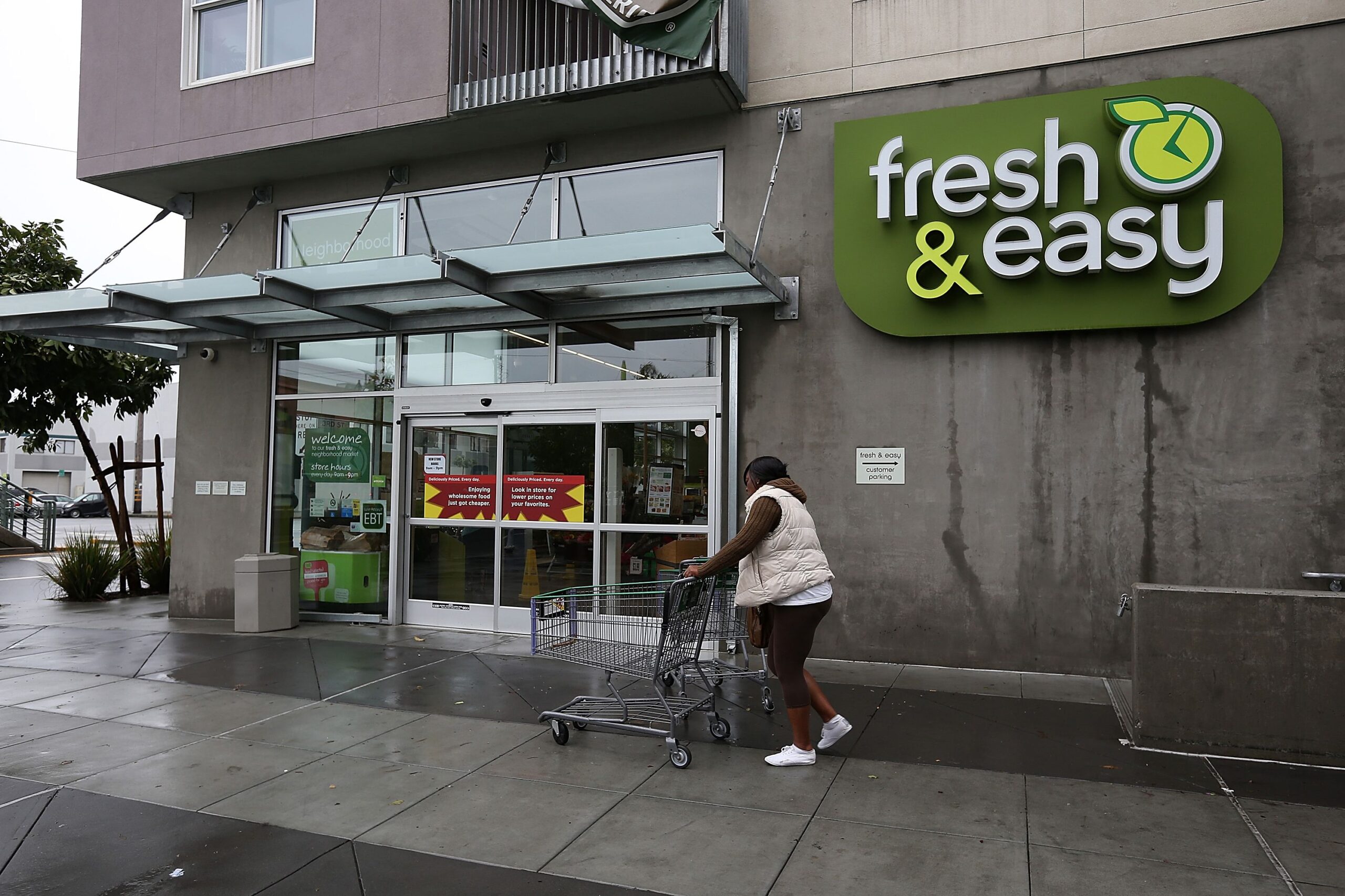 Front exterior of Fresh & Easy, San Francisco, 2012, woman is walking towards the glass doors with a shopping cart, sidewalk in the foreground, on a rainy day