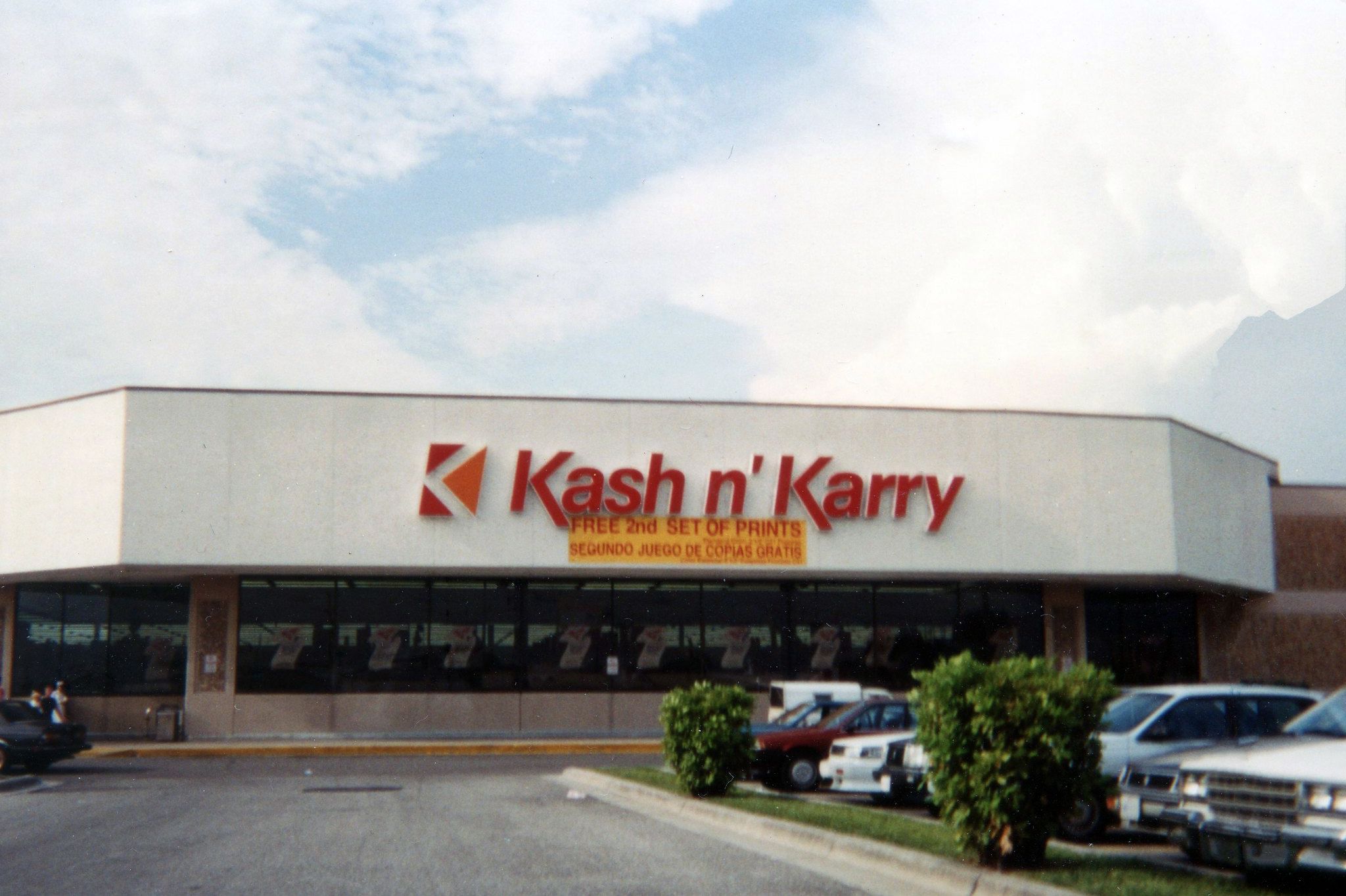 Front exterior of Kash n' Karry, Circa 1993, parking lot in the foreground, white top of building with brown bottom portion, a blue sky with white clouds