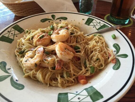 A plate of shrimp scampi pasta with angel hair noodles, shrimp, diced tomatoes, and green onions, garnished with black pepper&mdash;just like you'd find at Italian chain restaurants in America&mdash;served on a decorative plate with a fork.
