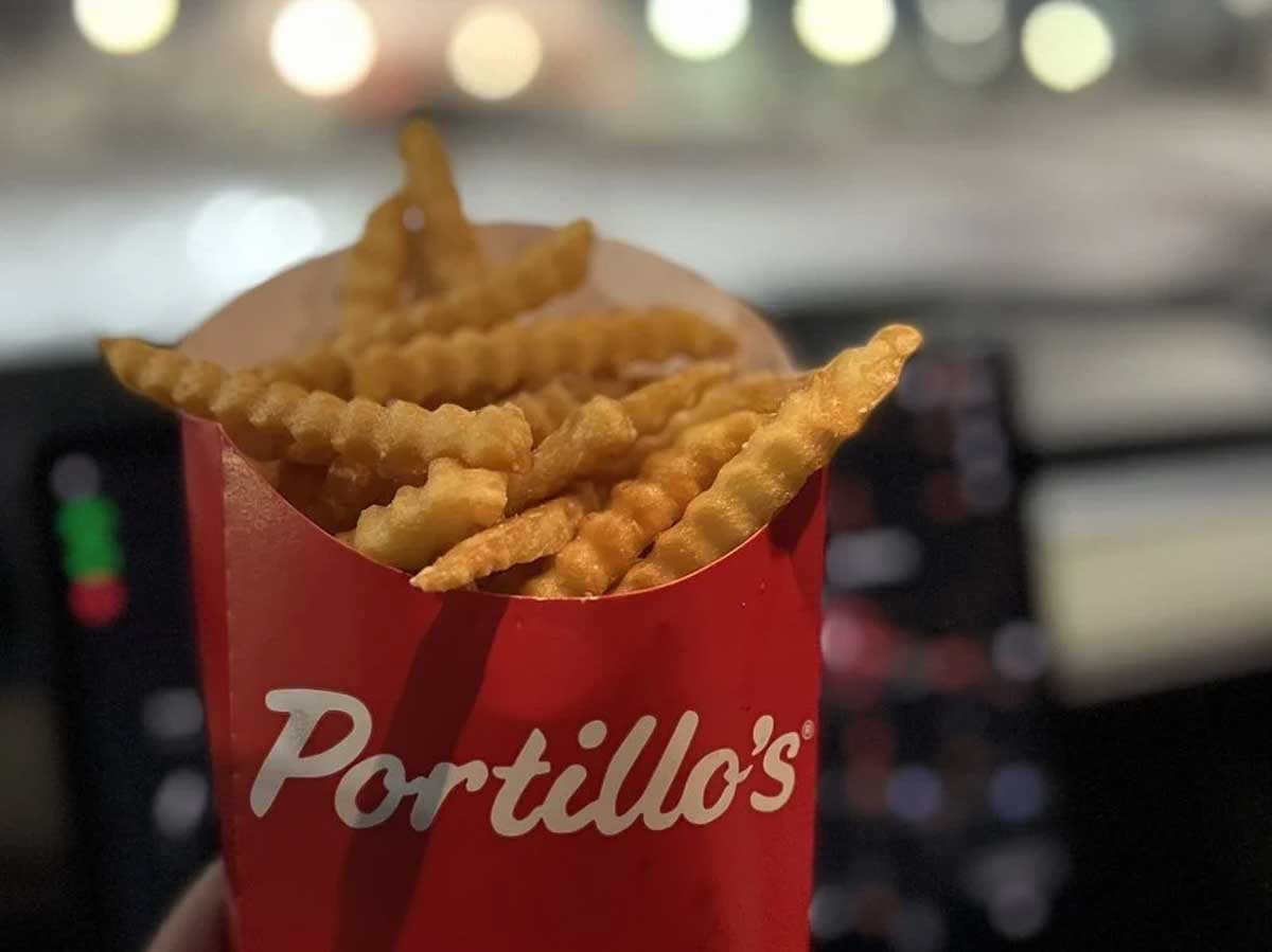 A red Portillo’s container filled with crinkle-cut beef tallow fries is held up against a blurred background, possibly inside a car at night with lights shining outside.