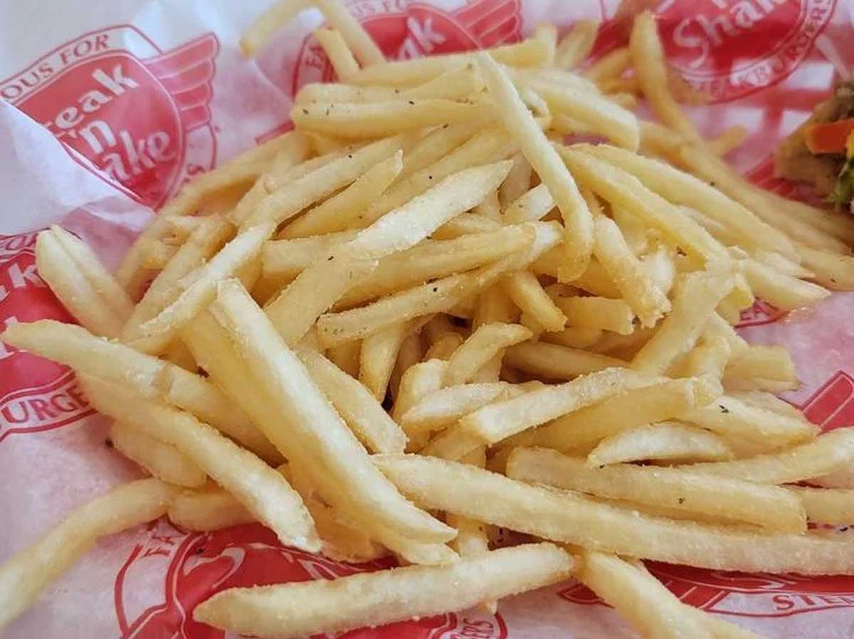 A pile of golden, crispy beef tallow fries rests on red and white branded paper from a restaurant, with a small portion of pickles visible in the corner.