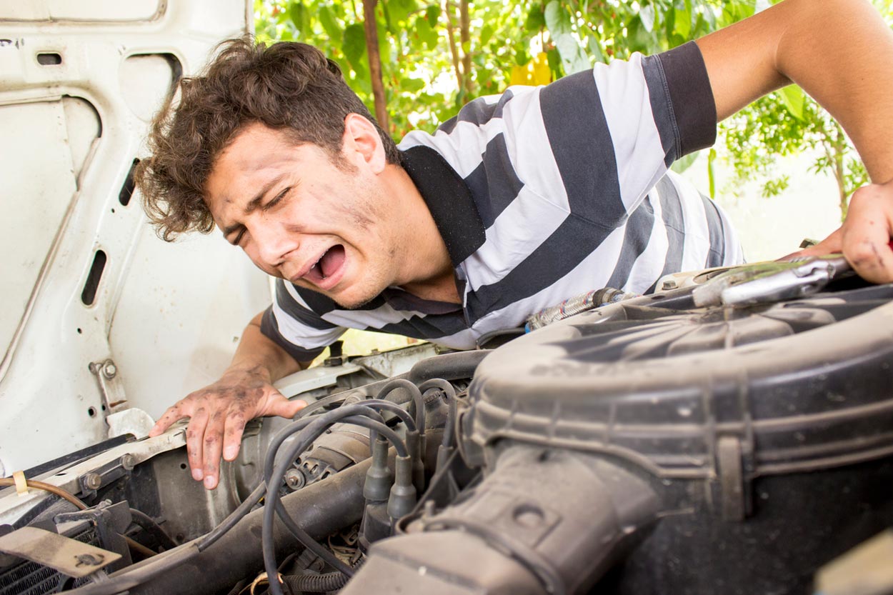 A young man with a distressed expression and dirty hands is struggling to fix a car engine, leaning over the open hood while holding a wrench. Trees are visible in the background.