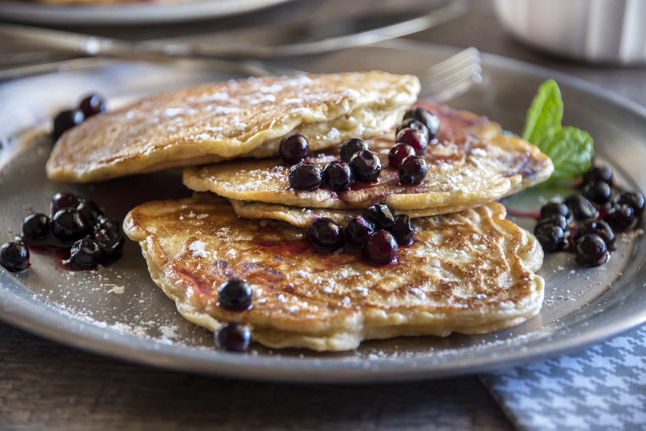 Rustic sourdough blueberry pancakes served on a pewter plate. The pancakes are sprinkled with powdered sugar and garnished with fresh mint. There is a second plate of pancakes in the background. Focus is in the center of the frame.