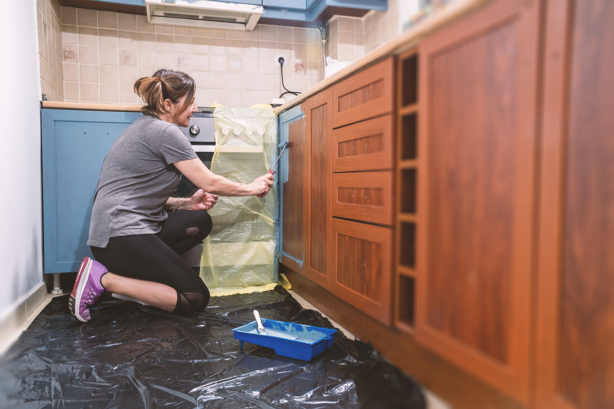 Mid age woman painting her old brown kitchen to blue modern color. Improving home is the main hobby for lockdown days.