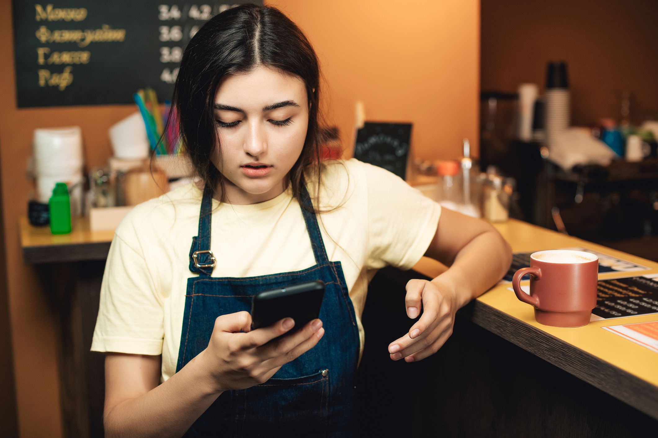 One cute woman working as bartender indoors cafe. Girl is using mobile phone and drink a coffee