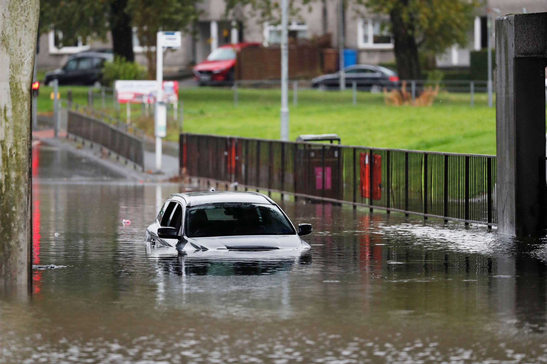 A red car is partially submerged in a flooded street, its wheels and bottom half of the car visible