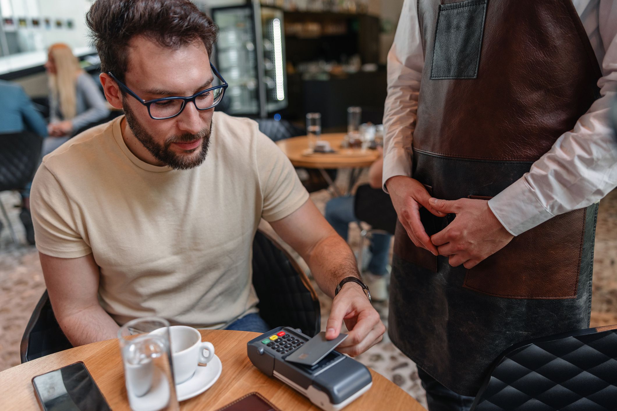 Attractive young adult Caucasian male paying with a contactless credit card at a coffee shop. The man has put his card on the POS terminal in order to pay. A waiter in an apron is standing next to him and waiting for the transaction to go through.