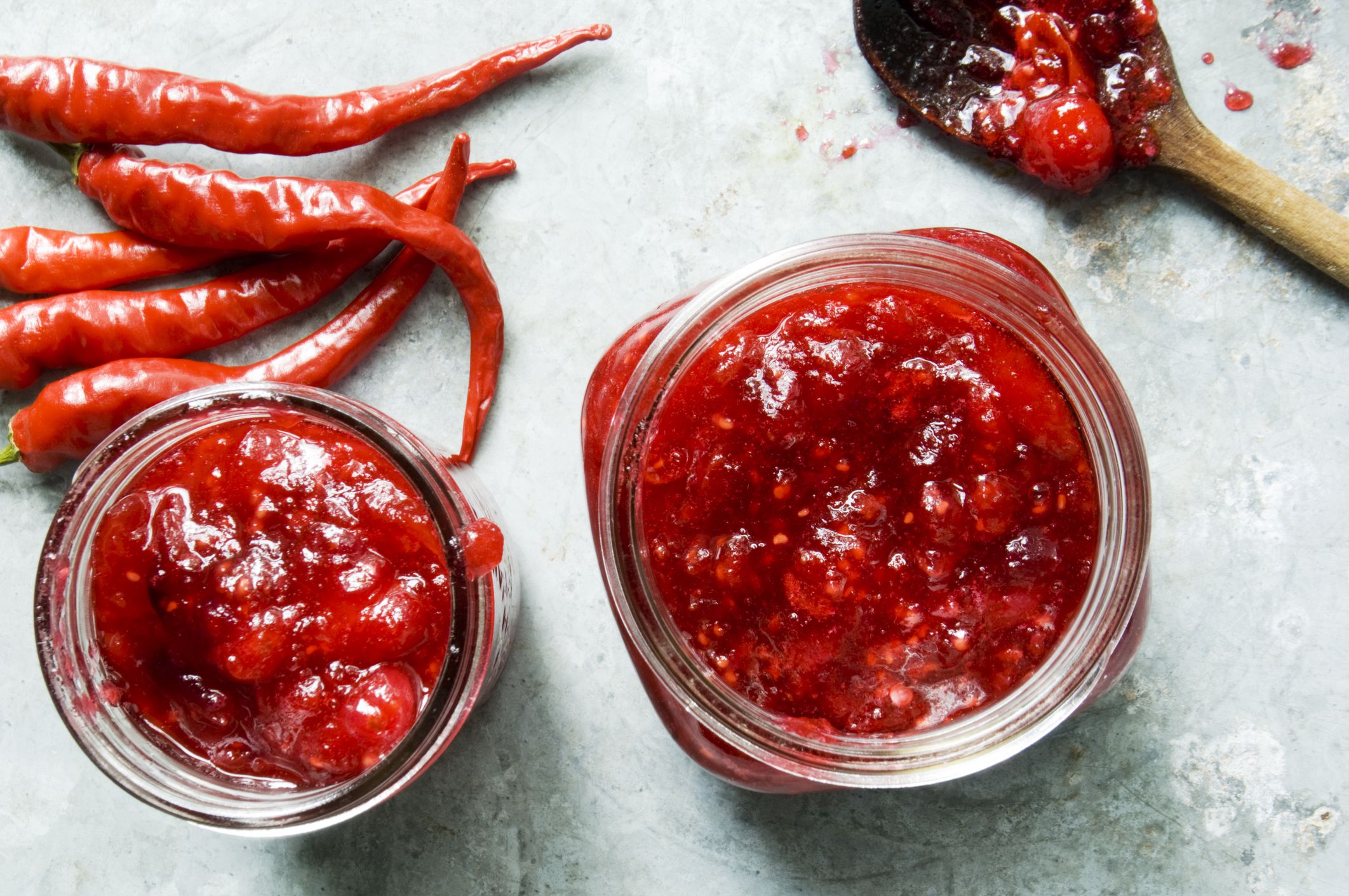 Two jam jars filled with cranberry and red chile jam are ready to be processed for storing. The image shot directly from above using natural light shows unused red chiles, a wooden spoon and jars of jam.