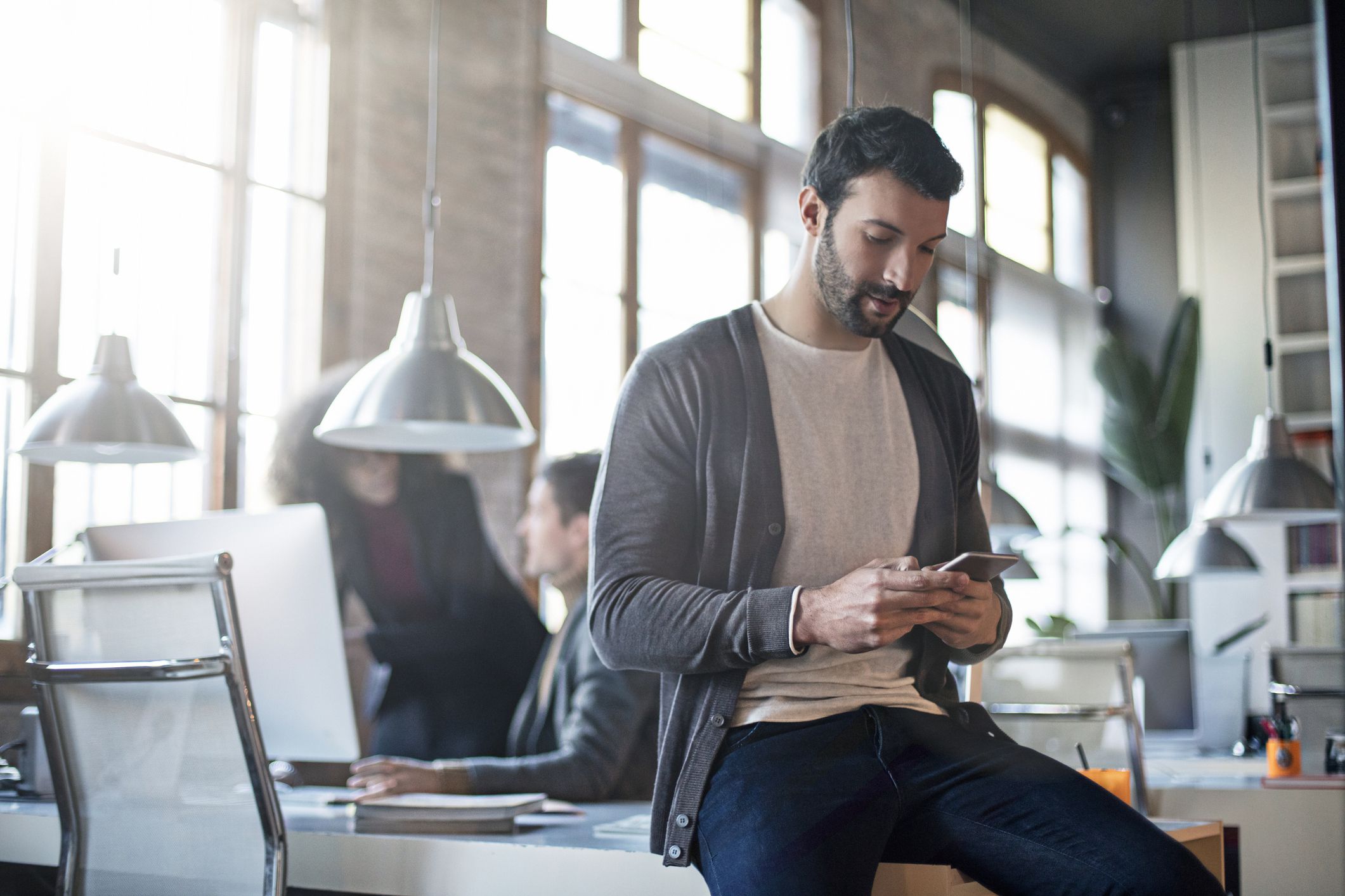 Businessman checking the mobile phone sitting on his desk, colleagues working in the background.