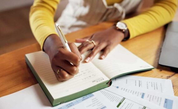 A person wearing a yellow sweater writes New Year's resolutions in an open notebook with a pen, while papers and documents about ways to save money are spread out on the table nearby.