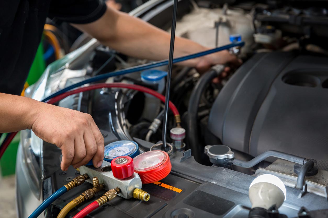A mechanic uses diagnostic gauges and hoses to service a car's air conditioning system under the open hood, adjusting controls and checking engine components.