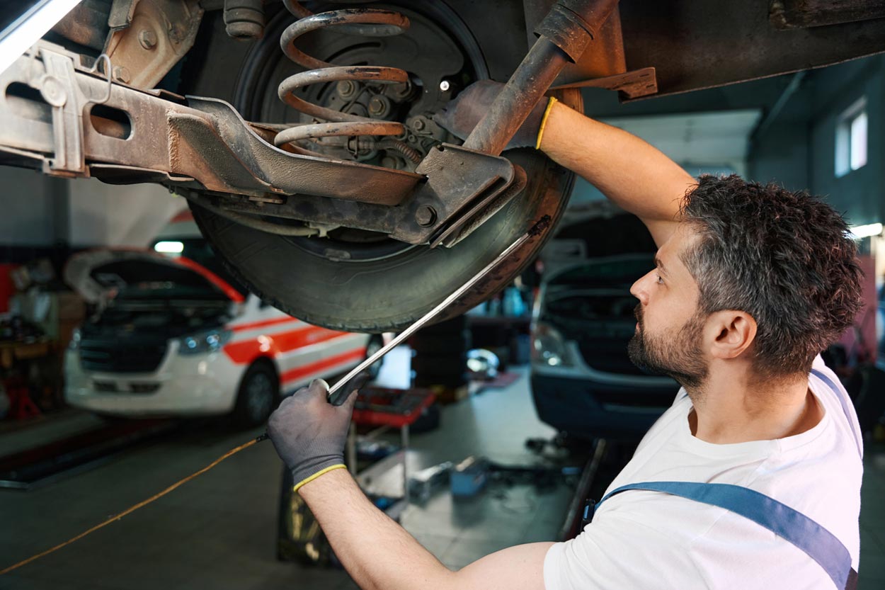 A mechanic wearing gloves works underneath a raised vehicle, using a wrench to adjust the suspension. Other cars with open hoods are visible in the background inside the auto repair shop.