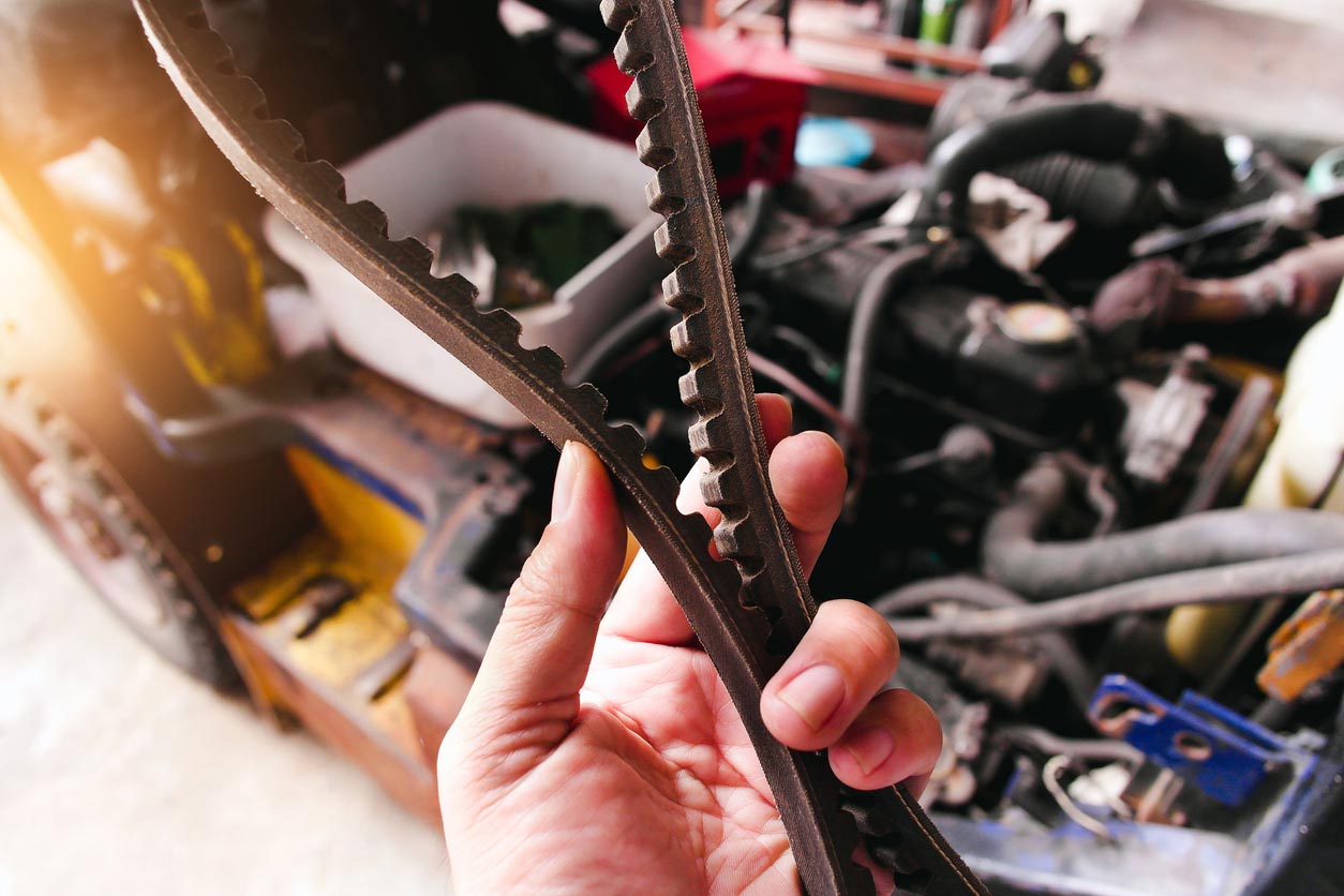 A hand holds a worn, damaged serpentine belt in front of an open car engine, highlighting automotive repair or maintenance work.