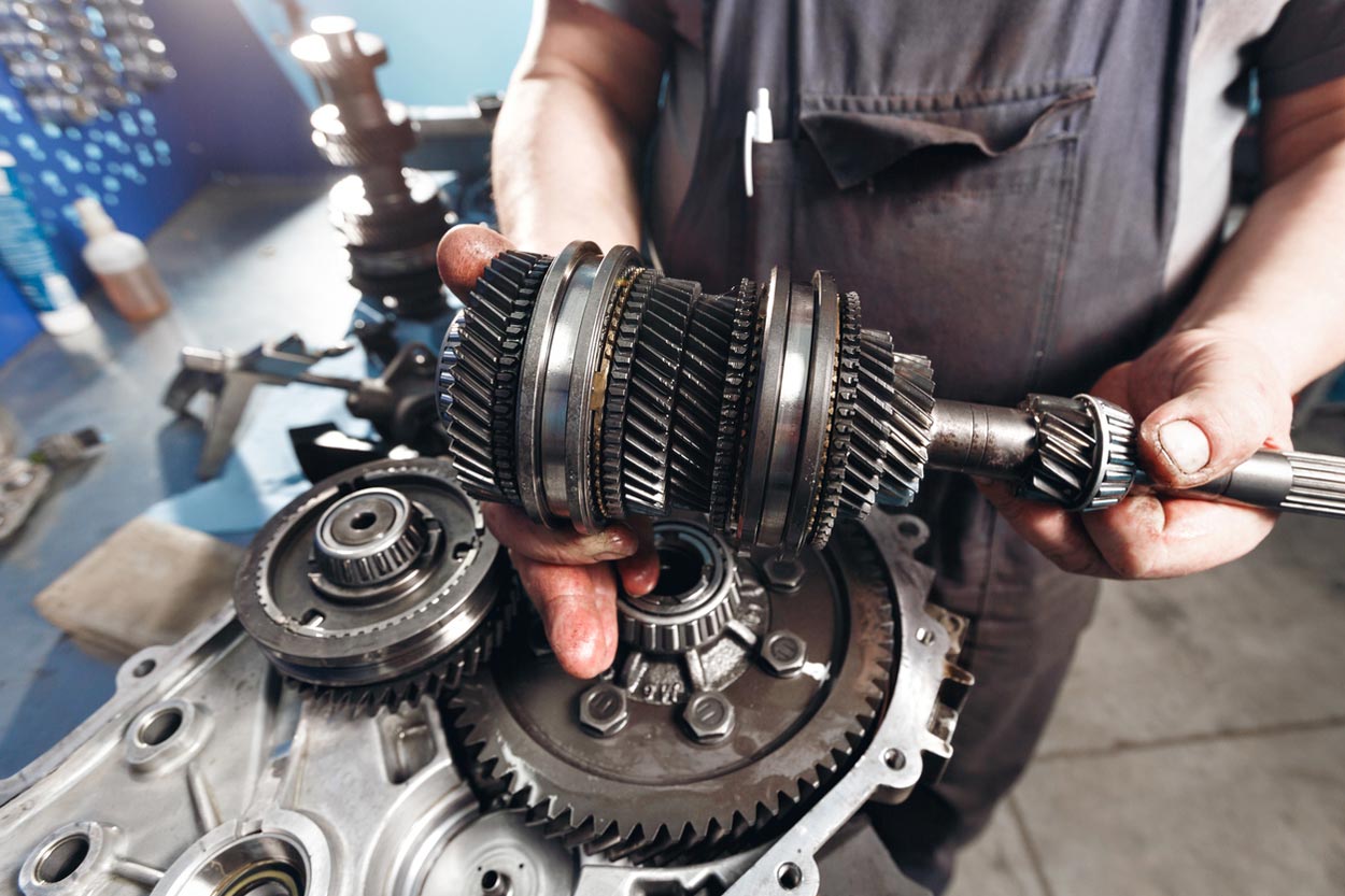 A mechanic wearing overalls holds a complex transmission gear assembly with both hands, surrounded by other metal engine components on a workbench in a workshop.