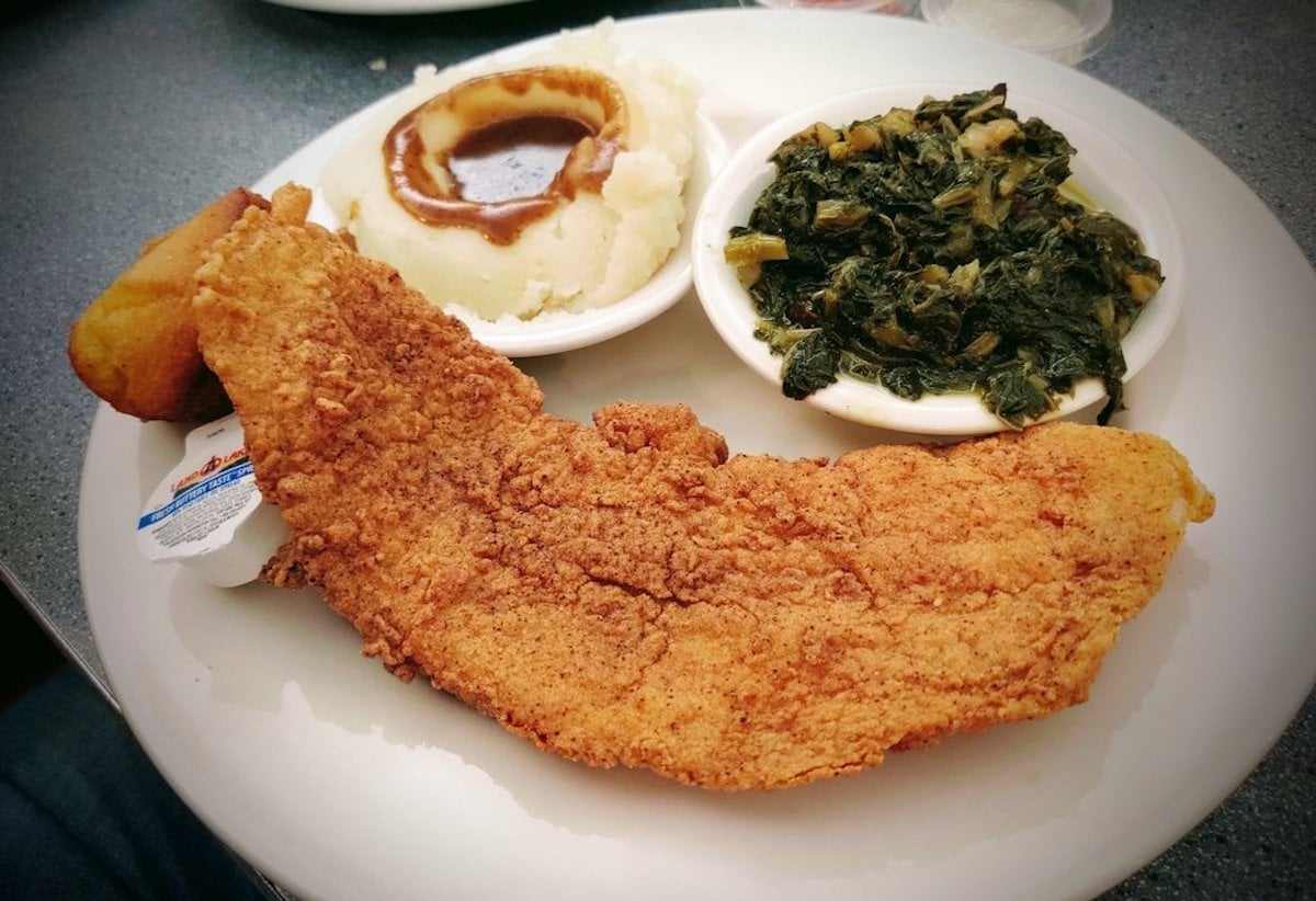 A plate of fried catfish, mashed potatoes with brown gravy, cooked greens, and a small cornbread muffin. The food is served on a white plate with a small container of tartar sauce.