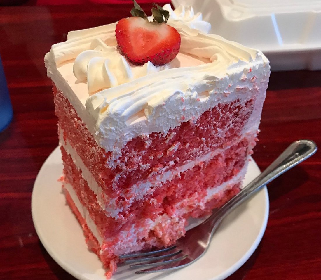 A slice of pink layered cake with white frosting and a sliced strawberry on top, served on a white plate with a fork.