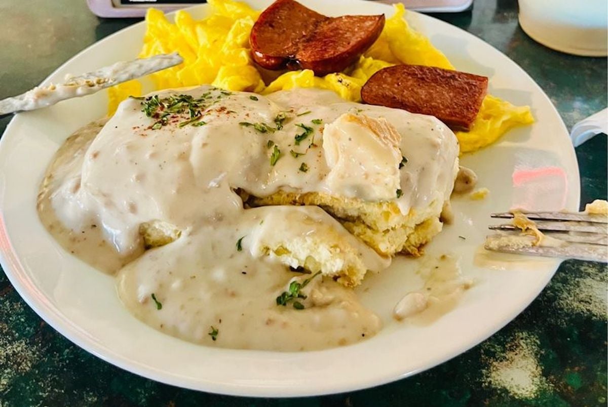 A plate with scrambled eggs, two slices of sausage, and a biscuit topped with creamy gravy and herbs. A fork and butter knife rest on the plate.