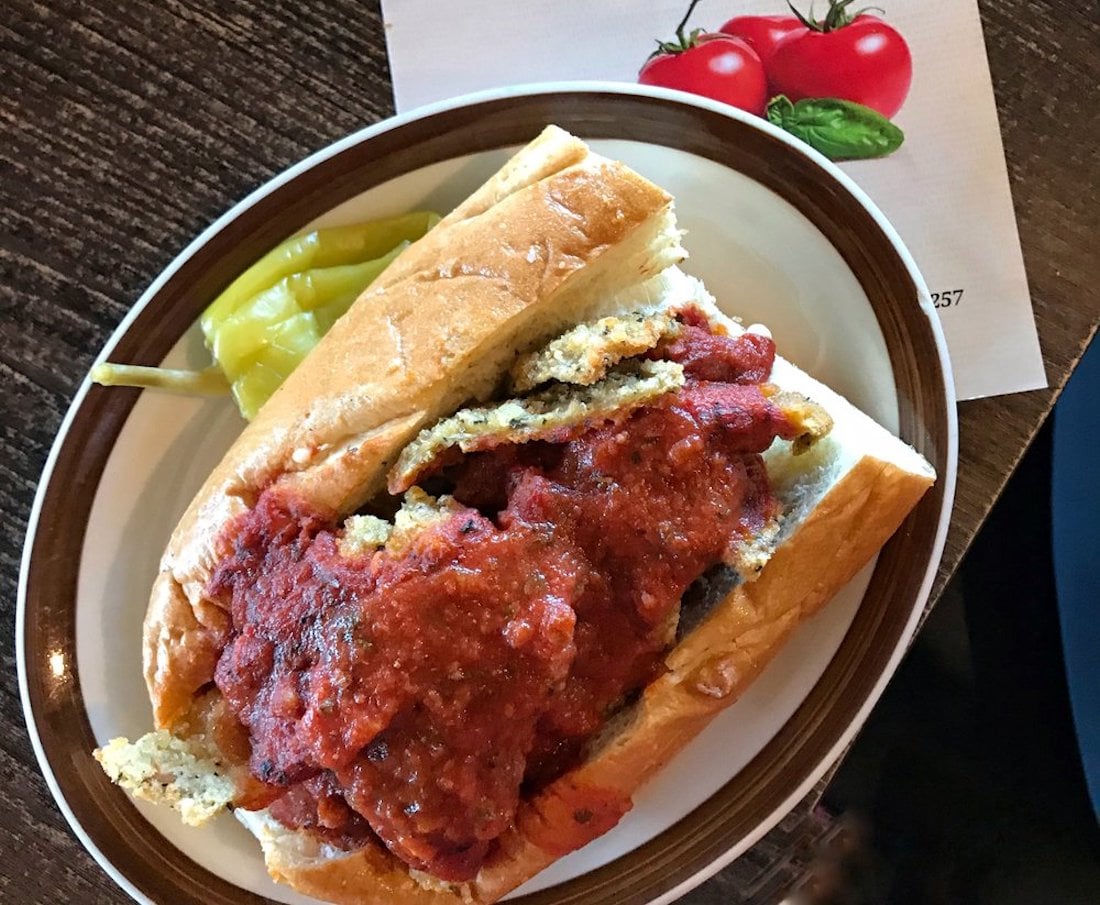 A sandwich roll filled with breaded cutlets and topped with marinara sauce sits on a plate beside a pickled pepper. A napkin with two illustrated tomatoes is partially visible in the background.