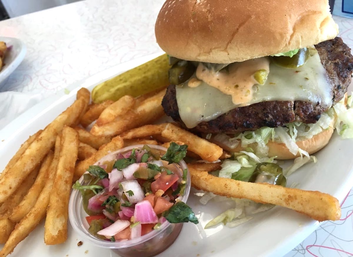 A cheeseburger with lettuce, jalapeños, and sauce on a bun, served with seasoned fries, a pickle spear, and a small cup of diced tomatoes, onions, and herbs on a white plate.