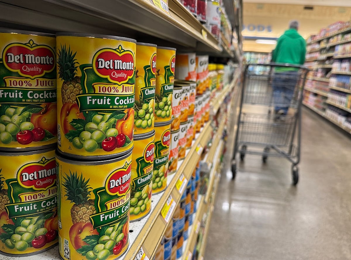 Cans of Del Monte fruit cocktail are stacked on a grocery store shelf. In the background, a person pushes a shopping cart down the aisle. The scene is brightly lit and focused on the cans.