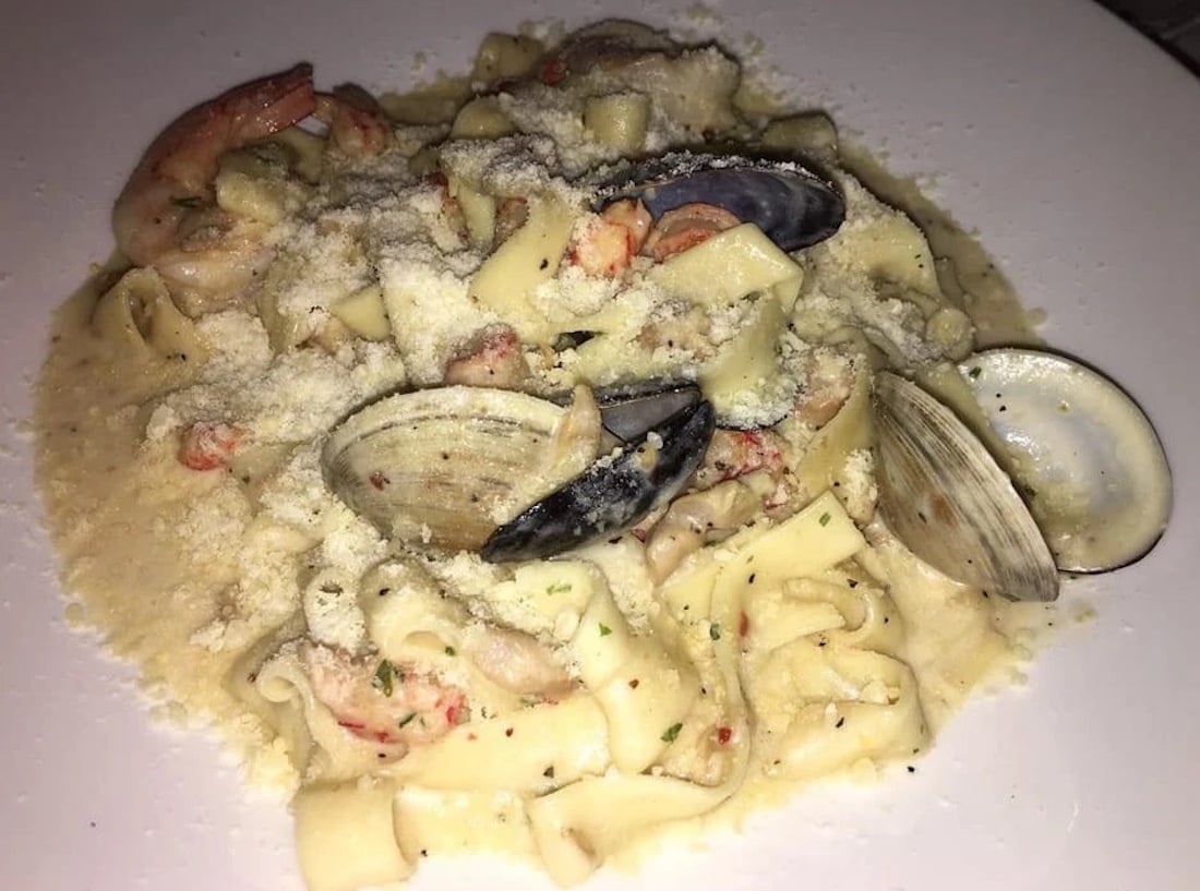 A plate of creamy pasta topped with grated cheese, mixed seafood including shrimp, clams, and possibly pieces of lobster, served on a white dish.