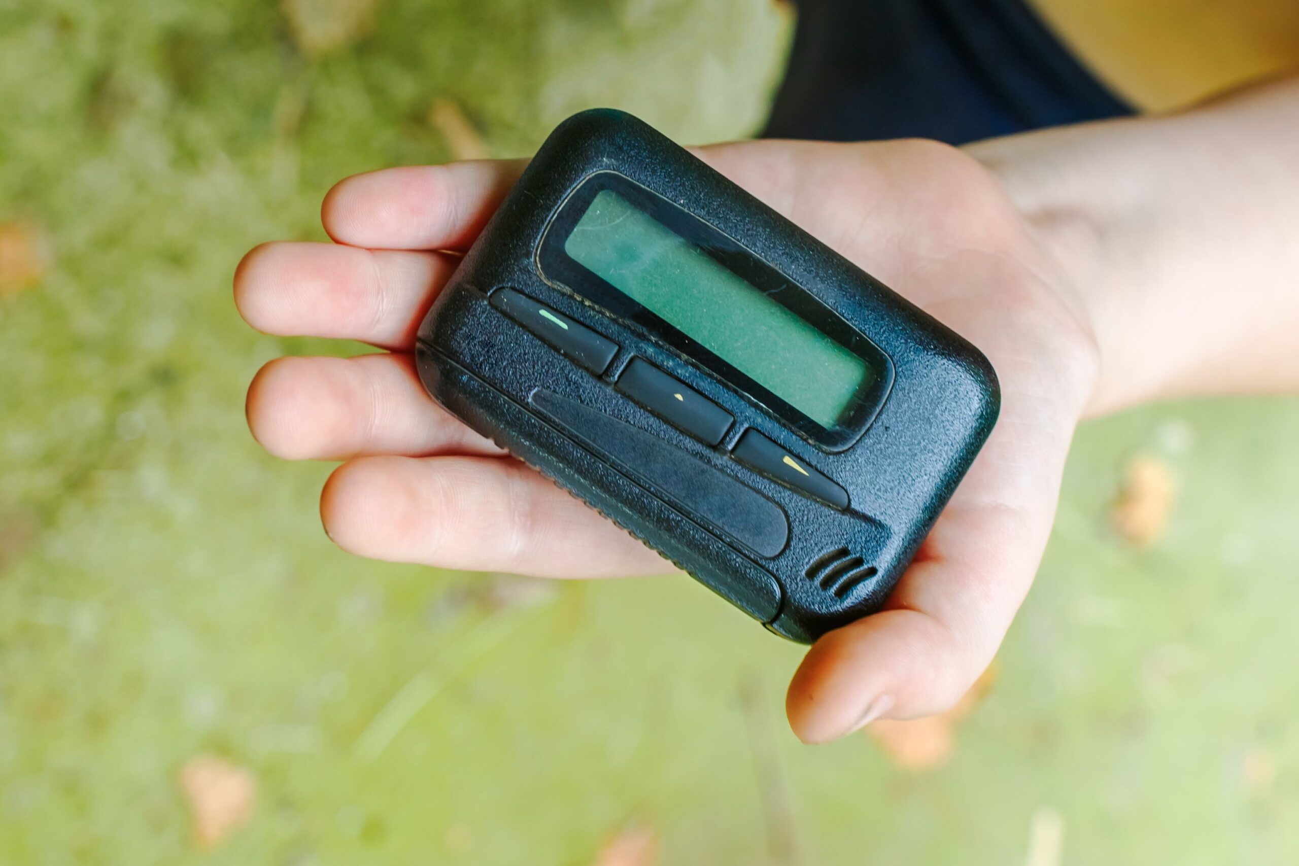 Looking from top, closeup of female hand holding vintage pager with a blurred background of a green floor with leaves, outside