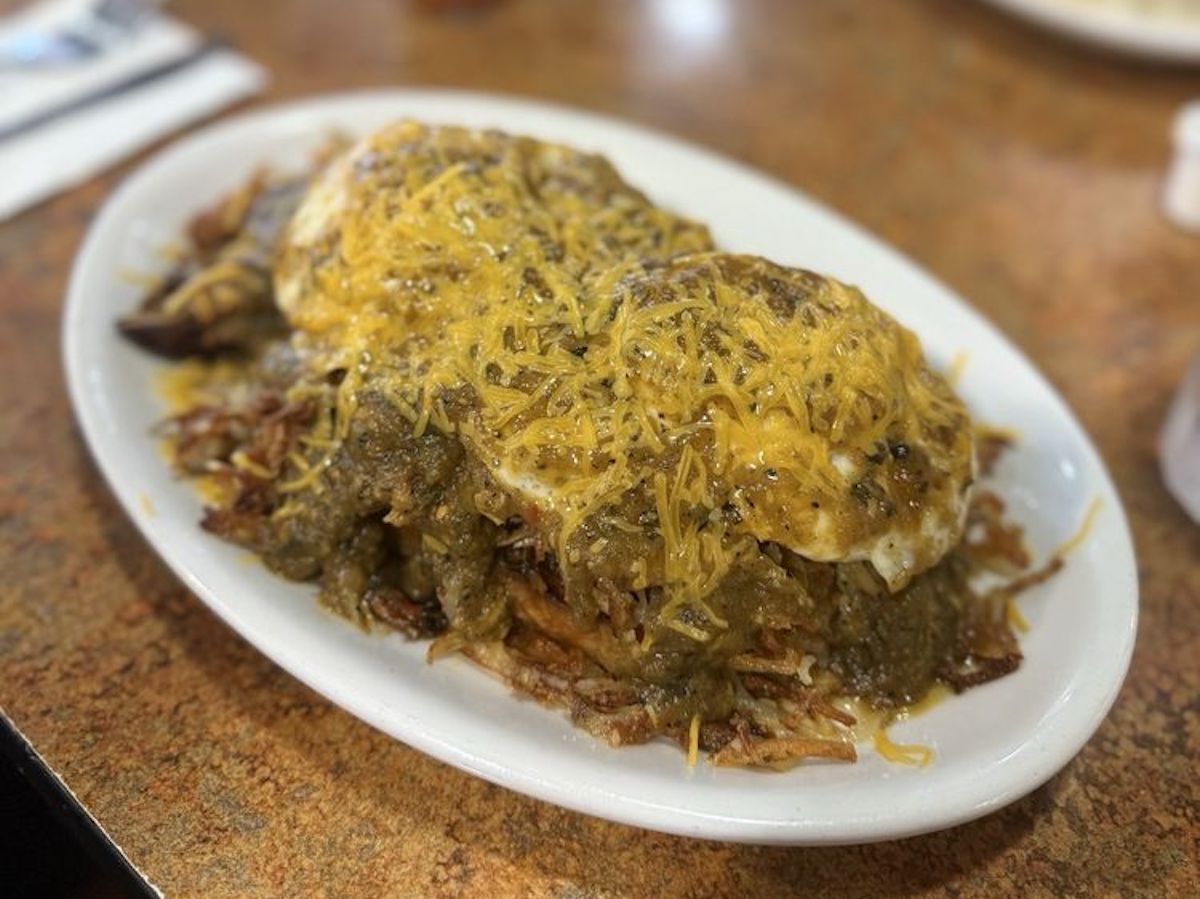 A white oval plate filled with crispy hash browns topped with eggs, shredded cheddar cheese, and green chili sauce, served on a brown table.