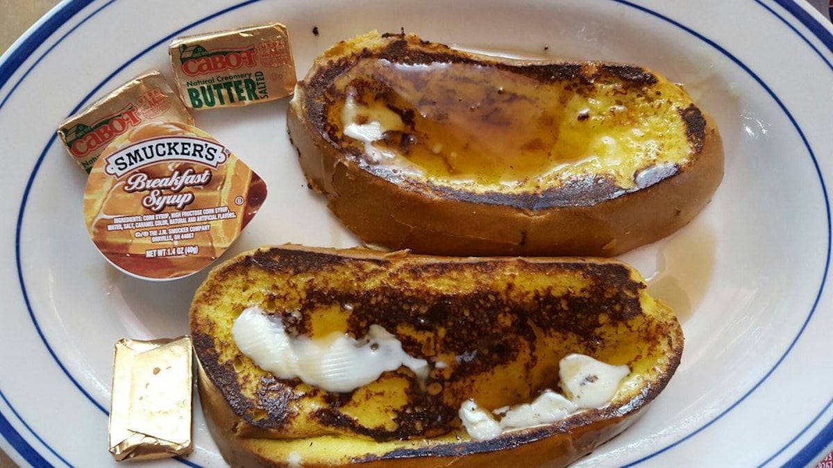 Two slices of French toast with melted butter and syrup on a white plate, served with packets of Cabot butter and Smucker’s breakfast syrup.