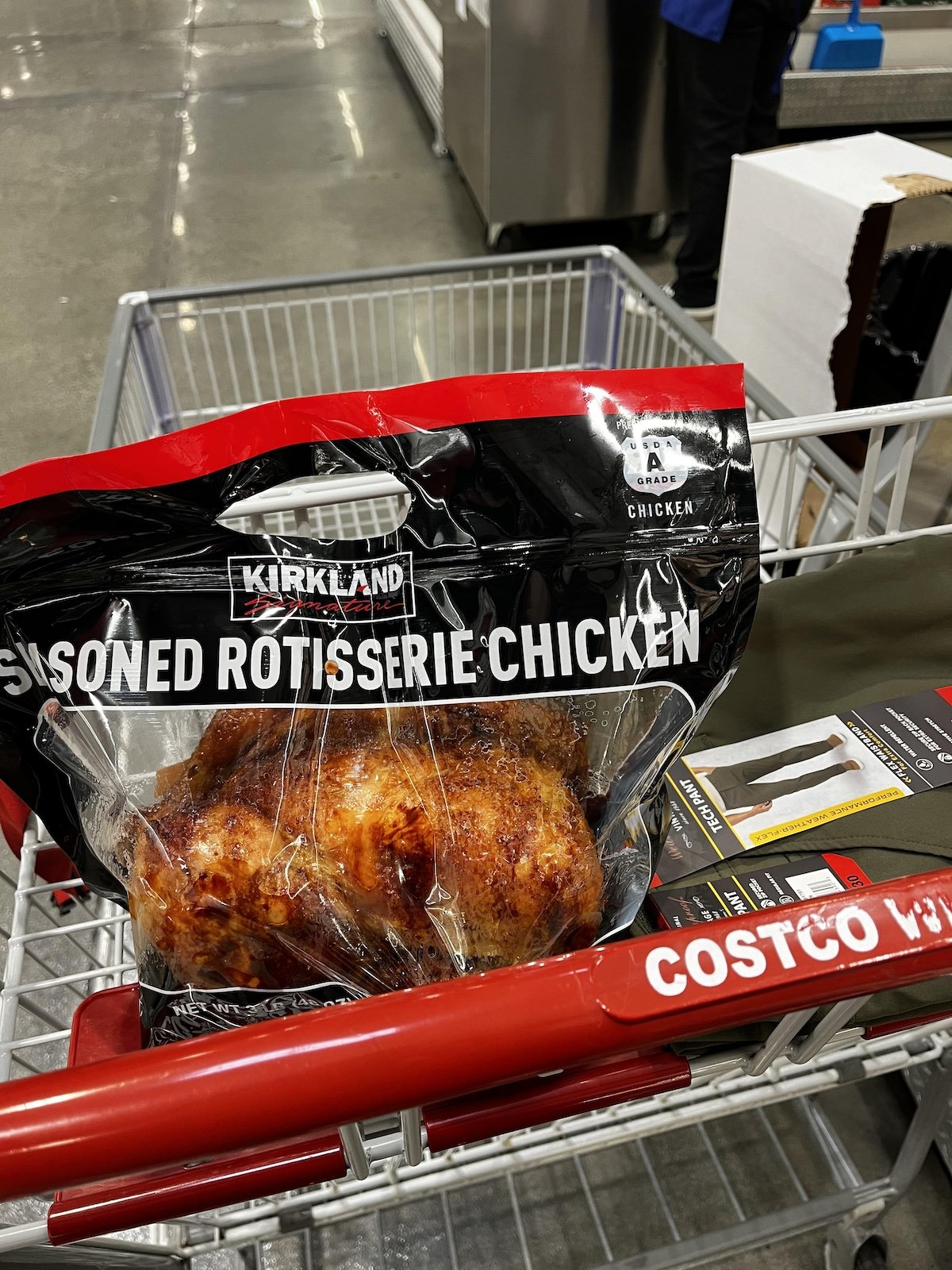A Kirkland Signature seasoned rotisserie chicken in a plastic bag sits in a Costco shopping cart along with a boxed item inside a warehouse store.