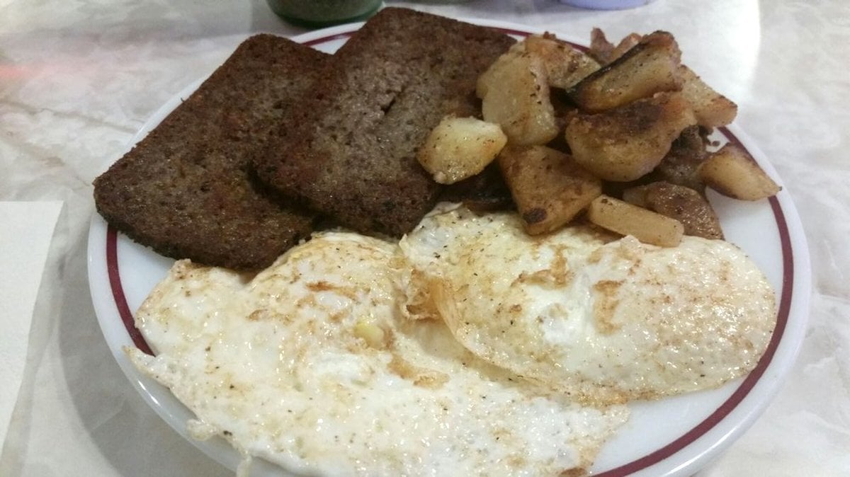 A plate with two fried eggs, three slices of cooked scrapple, and a serving of fried potatoes on a white plate with a red rim, set on a marble table.