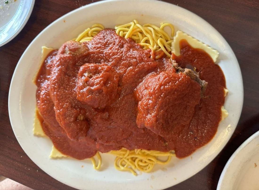 A plate of spaghetti and ravioli topped with meatballs and covered in marinara sauce, served on a white oval dish on a dark brown table.