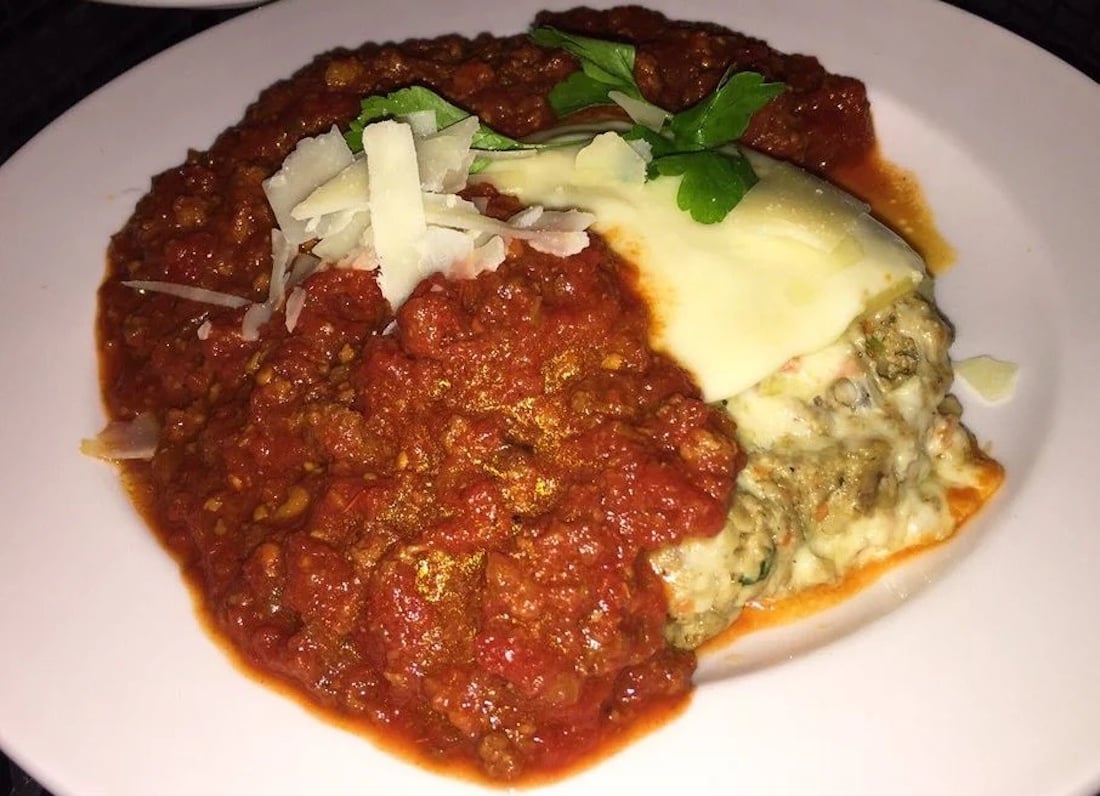 A slice of lasagna topped with melted cheese, meat sauce, and tomato sauce, garnished with fresh parsley and shaved parmesan, served on a white plate.