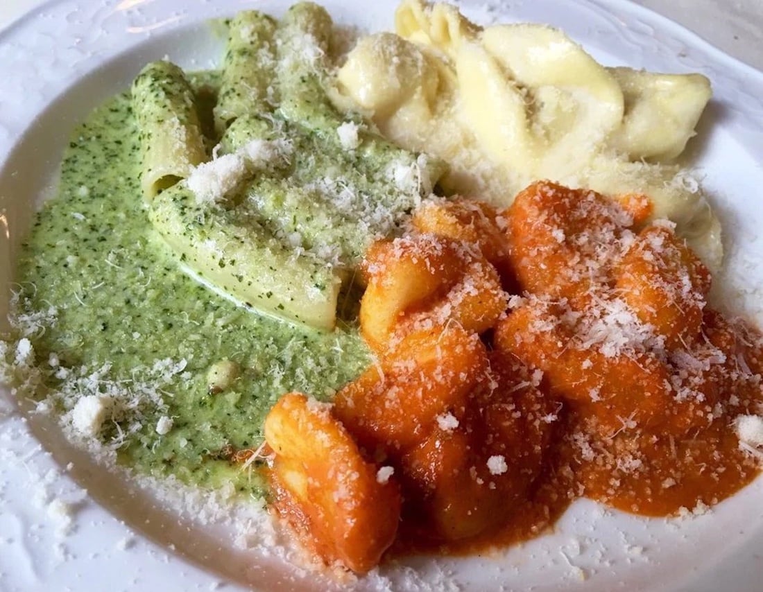 A white plate with three types of pasta: rigatoni in green pesto sauce, tortellini in cream sauce, and gnocchi in tomato sauce, all topped with grated parmesan cheese.