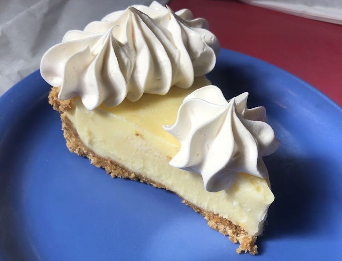 A slice of creamy yellow pie with a crumbly graham cracker crust, topped with two swirls of whipped cream, served on a blue plate.