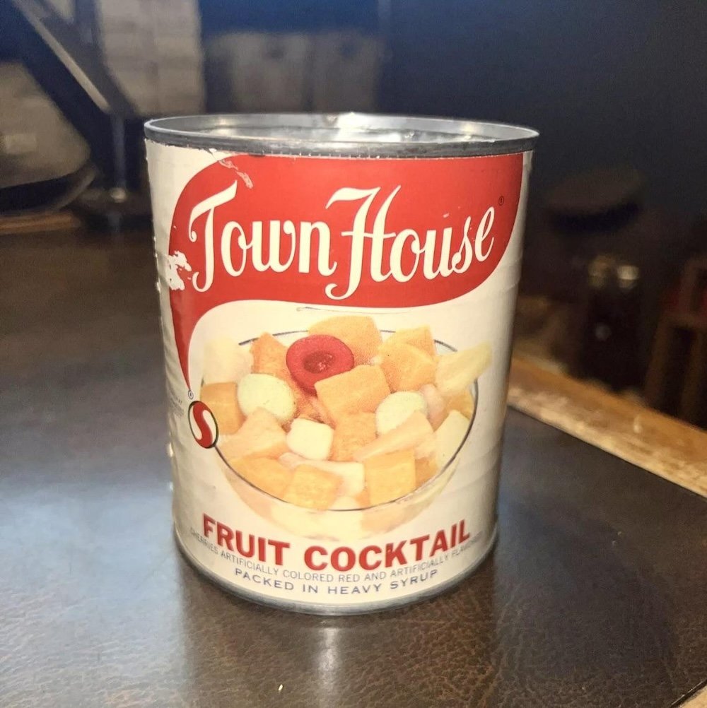 A can of Town House Fruit Cocktail featuring diced mixed fruit in heavy syrup, with a red and white label and an image of the fruit on the front, sitting on a dark surface.