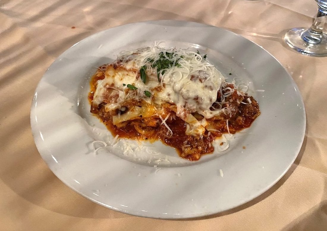 A plate of lasagna topped with melted cheese and grated parmesan, served on a white dish. The lasagna is layered with sauce and sits on a beige tablecloth next to a glass of water.