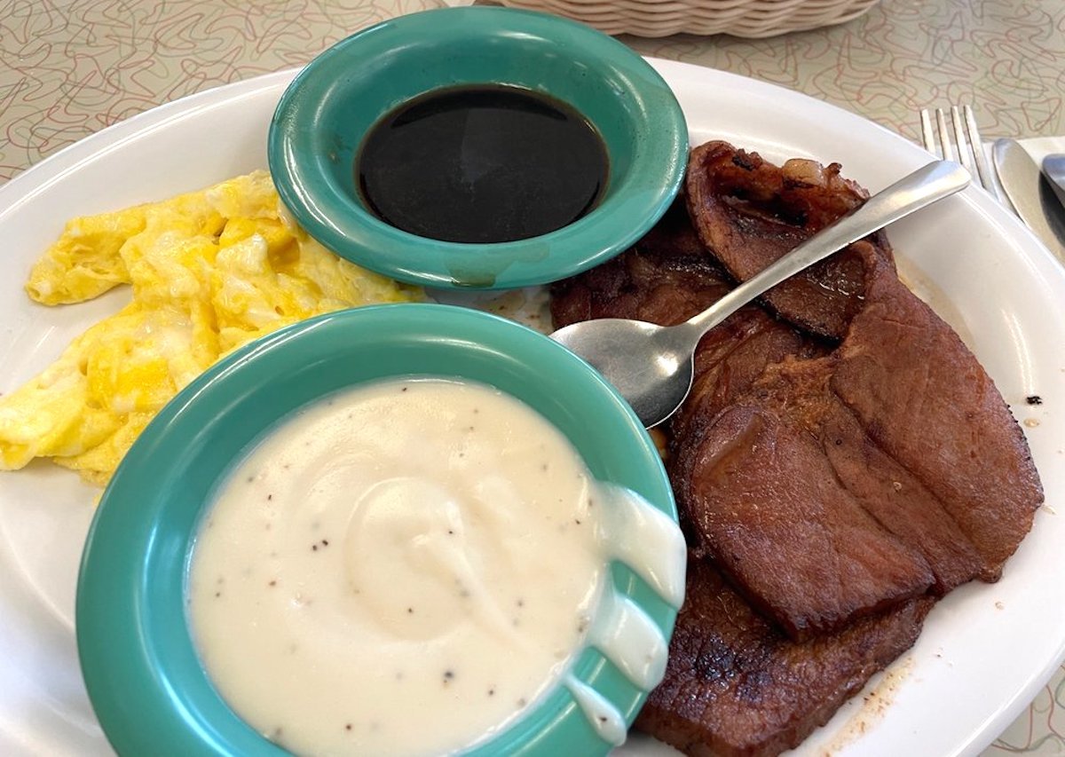 A plate with scrambled eggs, slices of ham, and two green bowls, one with brown gravy and one with white gravy, with a spoon resting on the ham.