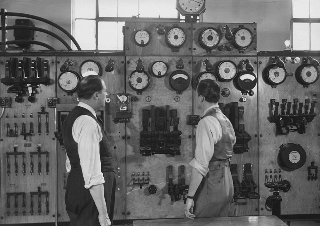 Two men stand in front of a console, inspecting the switches and dials, in a factory, USA, circa 1950.