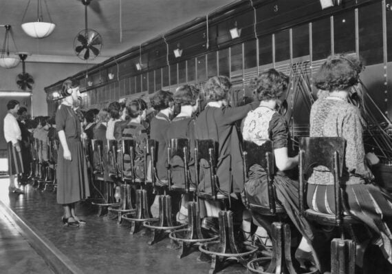 A row of female telephone operators sit on stools, wearing headsets and manually connecting calls at a large switchboard.
