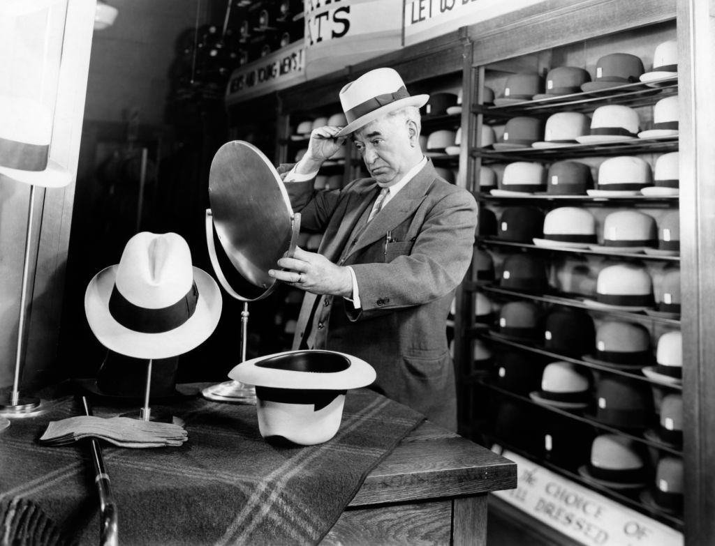 1940s SENIOR MAN TRYING ON HATS LOOKING IN MIRROR IN HAT STORE