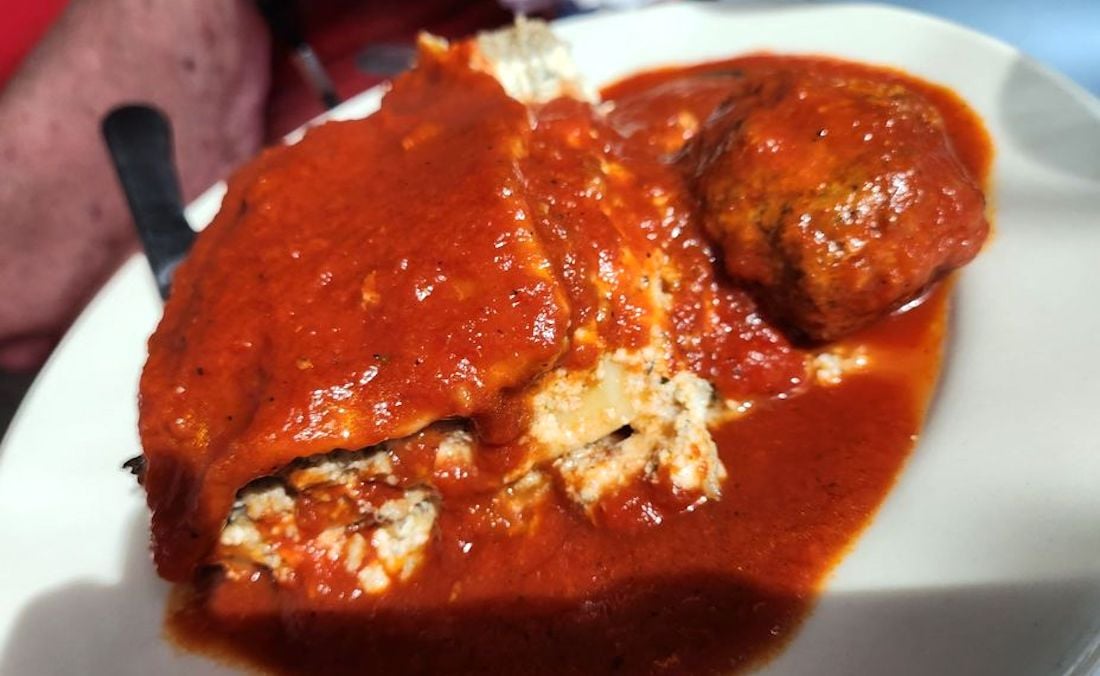 A close-up of a plate with lasagna and a meatball, both covered in a rich red tomato sauce.