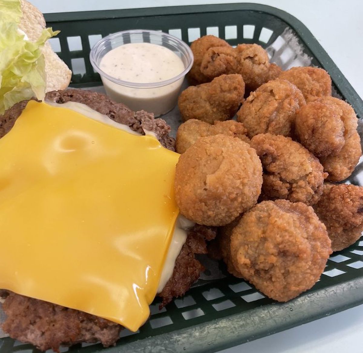 A basket featuring a cheeseburger patty with melted yellow cheese, a side of fried mushrooms, a cup of creamy dipping sauce, and a piece of lettuce on the side.