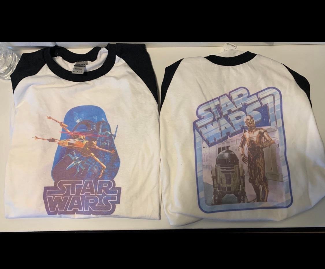 Two white and black raglan Star Wars T-shirts. The left shirt features Darth Vader, X-wing fighters, and the Star Wars logo. The right shirt shows C-3PO, R2-D2, and the Star Wars logo above them.