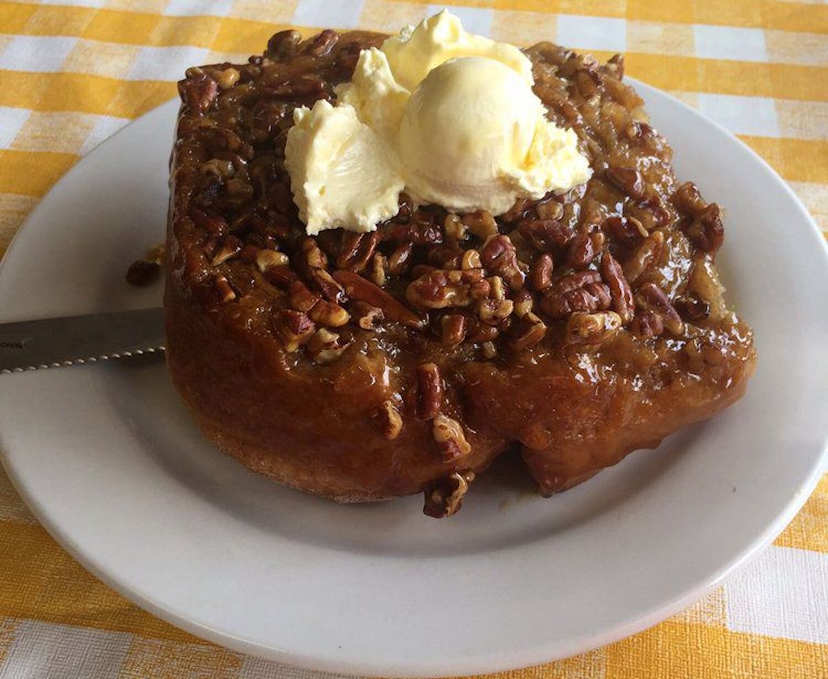 A large sticky bun topped with chopped pecans and two scoops of butter sits on a white plate. The plate rests on a yellow and white checkered tablecloth. A knife is partially visible under the bun.